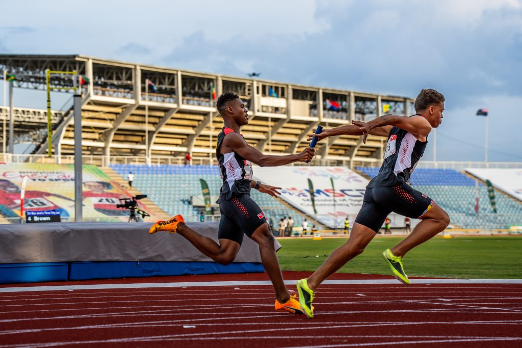 Team TTO athletes exchange the baton during the Boys 4x100m relay at the 2025 CARIFTA Games, with the stadium stands and flags visible in the background.