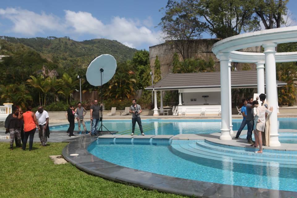 Vogue Italia production team sets up a fashion photoshoot at the Diplomatic Centre, Prime Minister's Residence in Trinidad. The crew arranges lighting and camera equipment beside a pool framed by white columns.