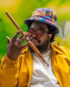 Rhys Thompson in colourful hat and yellow jacket playing a wooden drumstick.
