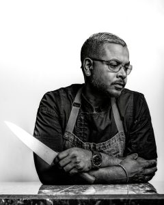 Chef Aaron Farrick holding a kitchen knife across his chest in a black and white portrait.