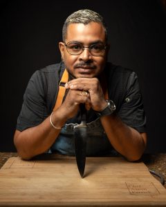 Chef Aaron Farrick leaning over a cutting board with hands clasped around a chef’s knife.