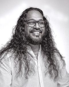 Robin Bawhanie smiling in a black-and-white portrait with long hair and glasses.