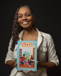 Aliya Drakes smiling while holding her book It Takes a Village.